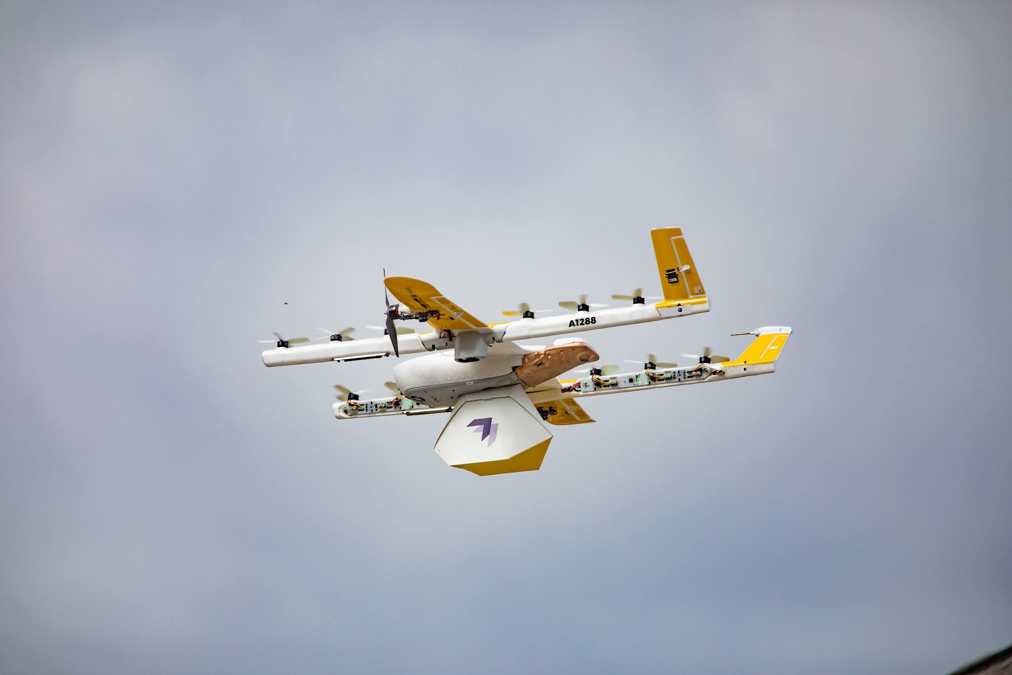 A yellow and white drone with multiple propellers is flying in a clear sky, carrying a package underneath, demonstrating modern drone delivery technology.