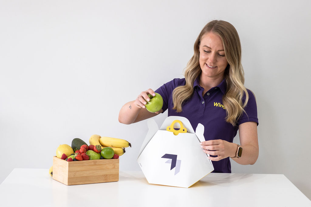 A person in a purple polo shirt is placing a green apple into a white, hexagonal food delivery box with a logo, next to a wooden crate filled with bananas, avocados, and strawberries on a white table.