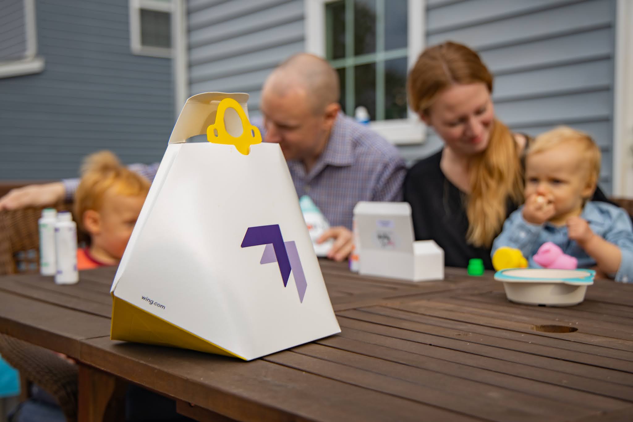 A family enjoys a vibrant outdoor gathering on a wooden table, featuring a focus on Wing's modern delivery container with a stylized purple arrow, ideal for highlighting outdoor dining and family meals.