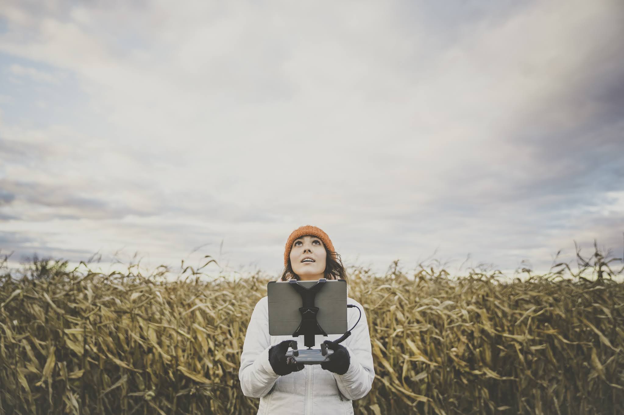 A person wearing a bright orange beanie and white jacket stands in a vast cornfield holding a drone controller, gazing upwards at the cloudy sky.
