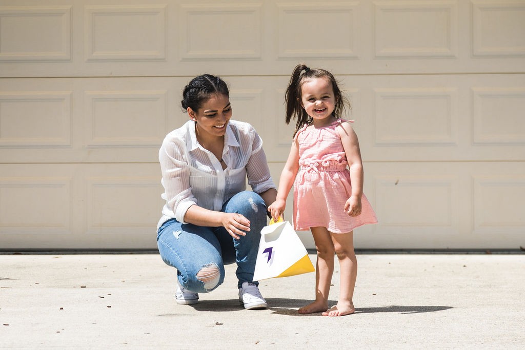 A woman crouches on a driveway in front of a garage door, smiling at a young girl in a peach dress holding a  中国168体彩网-实时官方认证 Delivery package.