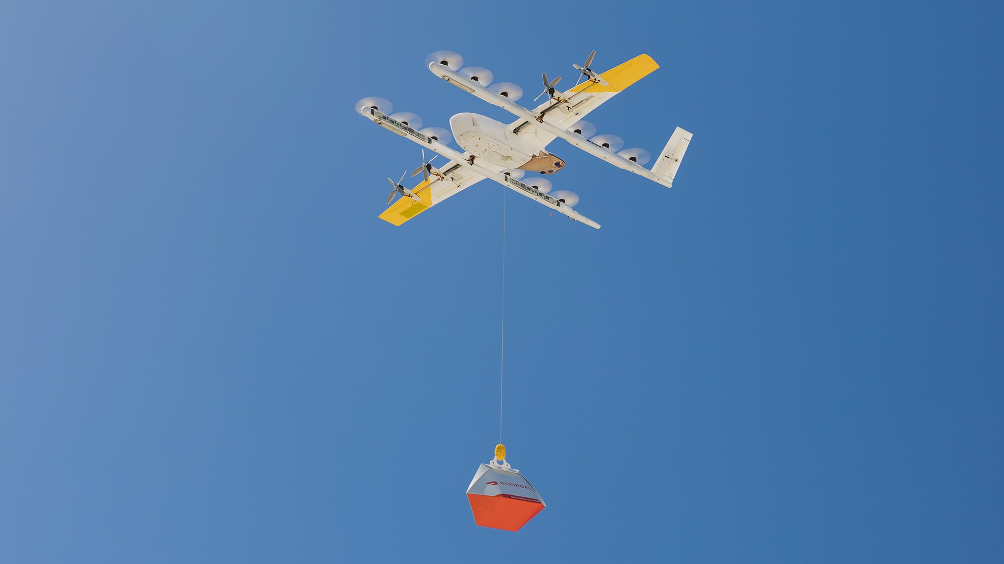 A white and yellow drone in flight against a clear blue sky; it is carrying a red and orange package, demonstrating modern delivery technology.