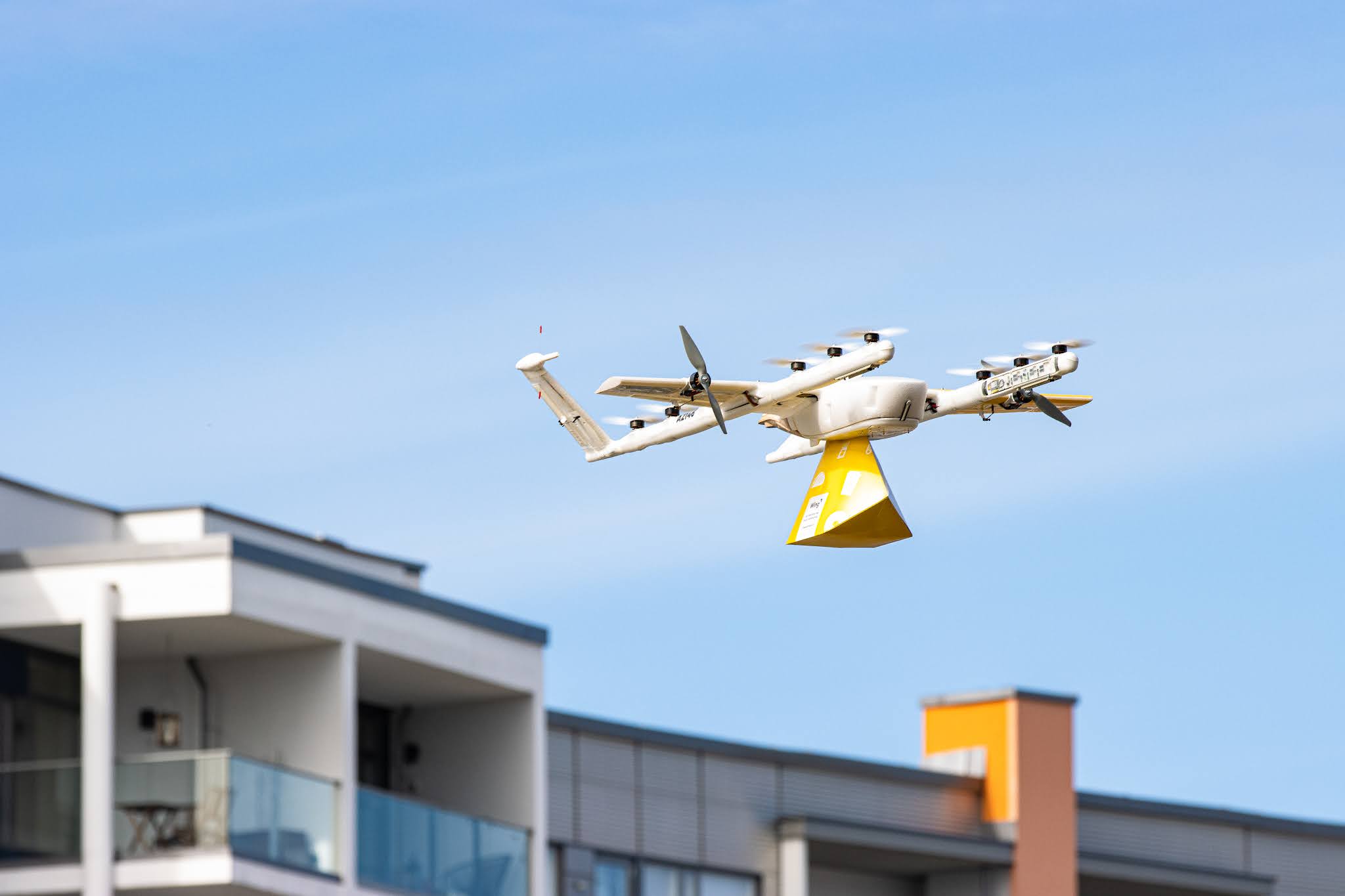 A delivery drone flies over a sprawling suburban neighborhood under a clear blue sky, showcasing modern technology in urban logistics and transport.