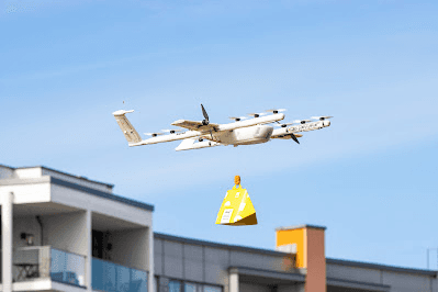A white delivery drone flies above urban apartments, carrying a yellow parcel beneath a clear blue sky.