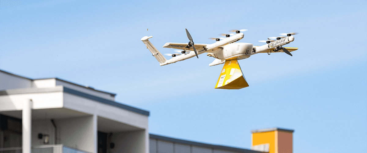 A delivery drone flies over a sprawling suburban neighborhood under a clear blue sky, showcasing modern technology in urban logistics and transport.