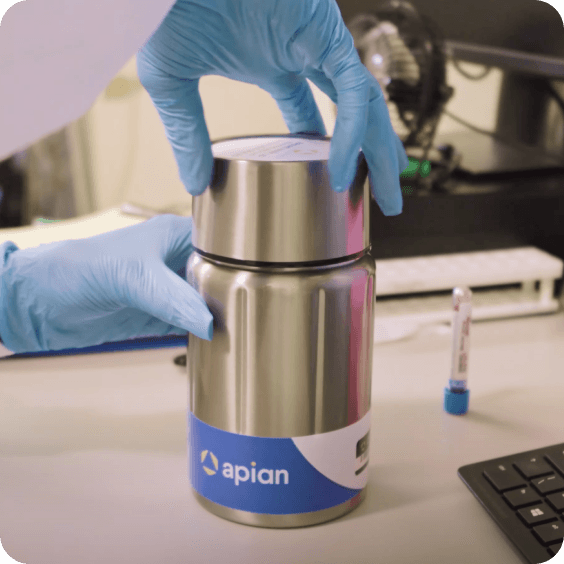 A pair of gloved hands securely closes a stainless steel container labeled "apian" on a laboratory desk, with laboratory equipment and a computer keyboard visible in the background.
